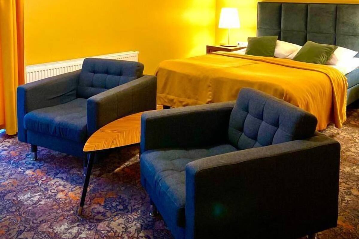 A hotel room with a yellow wall, a bed with a green headboard, two blue armchairs, a small table, and a lamp.