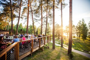 Un área de comedor al aire libre con mesas y sillas, rodeada de árboles altos y un espacio cubierto de césped.