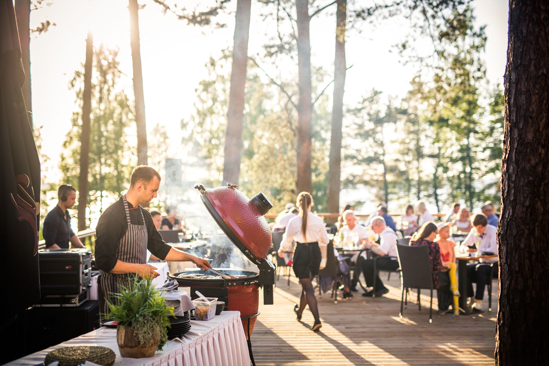 Un'area barbecue con griglia e utensili da cucina, circondata da ospiti che cenano a tavoli.