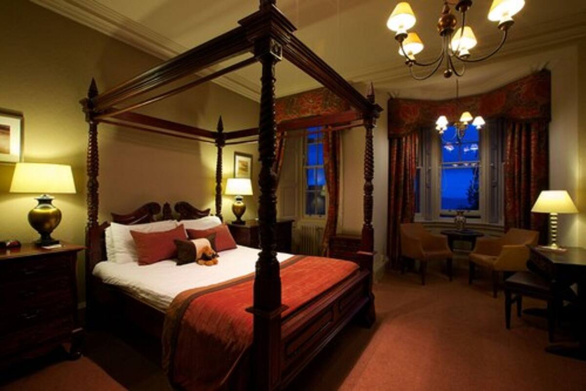 A four-poster bed with a red bedspread, two bedside tables with lamps, a chandelier, a window with curtains, and a small table with chairs in a hotel room.