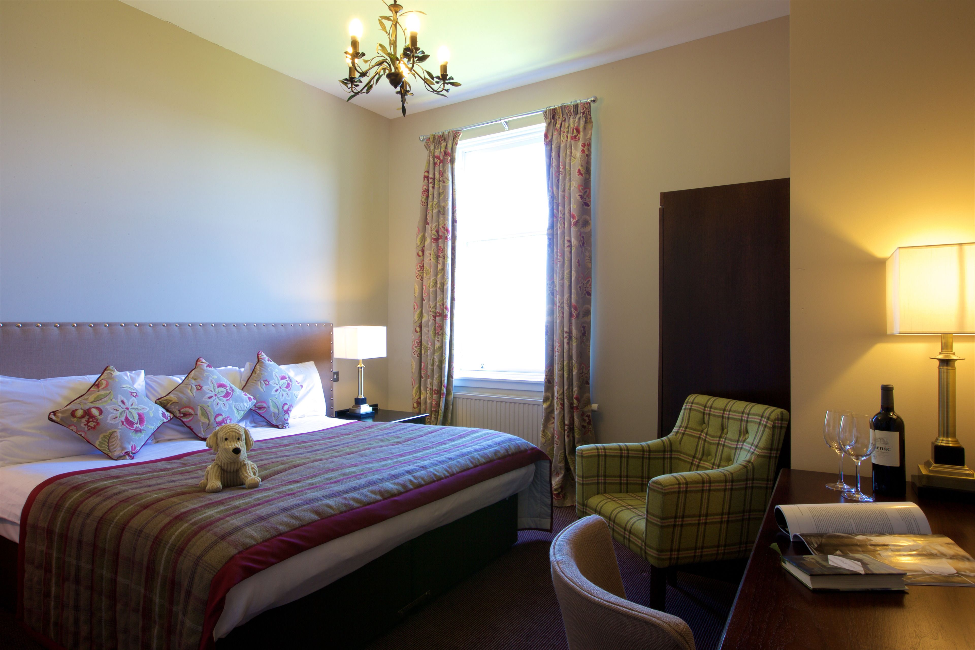 A hotel room with a bed, a chair, a lamp, a bottle of wine, and a window with curtains.