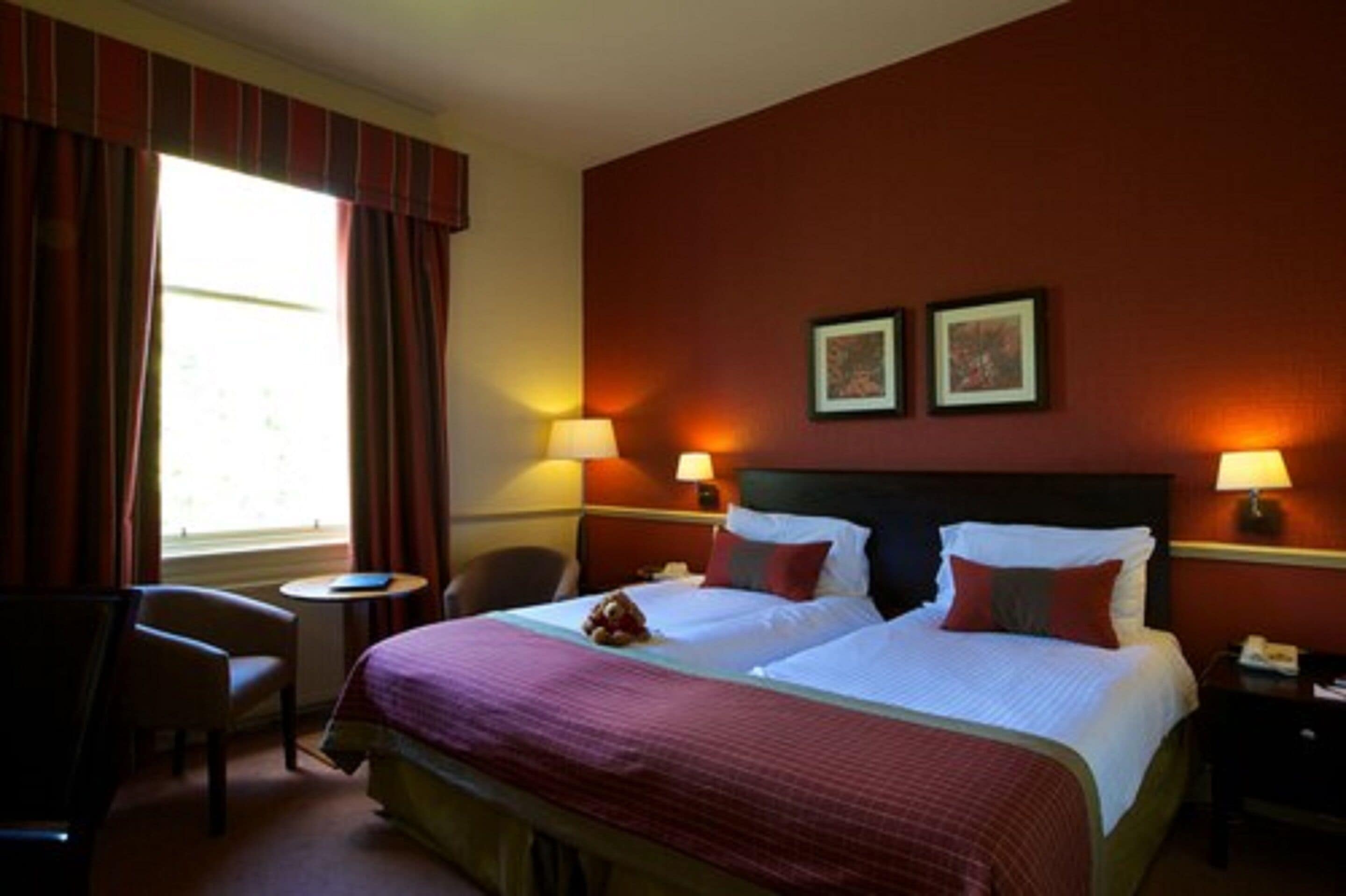 Quarto de hotel com duas camas, uma escrivaninha e uma cadeira. Há duas luminárias nas mesas de cabeceira e uma janela com cortinas.