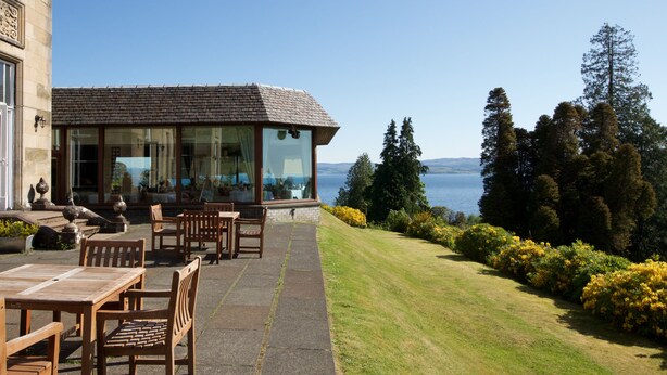 Un edificio con un patio amoblado con mesas y sillas de madera, vista al mar y un jardín arbolado con flores amarillas.