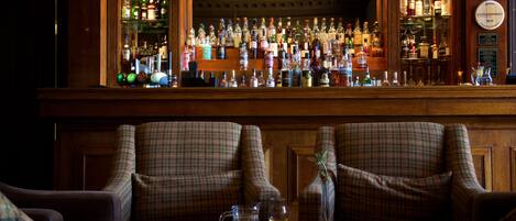 A bar area with a variety of liquor bottles, two plaid armchairs, and a small table with a candle and glasses.