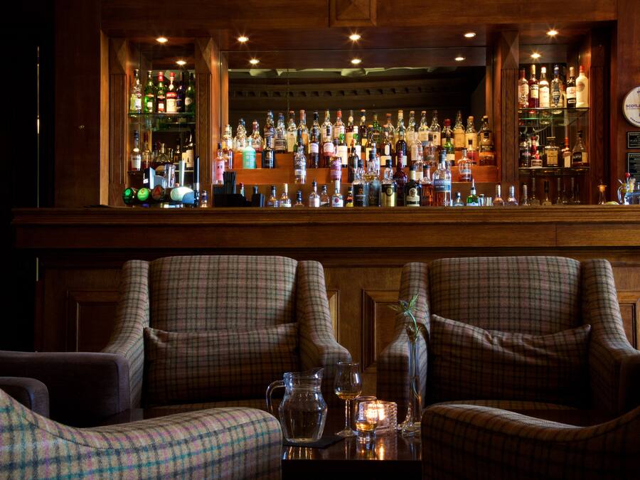 A bar area with a variety of liquor bottles, two plaid armchairs, and a small table with a candle and glasses.