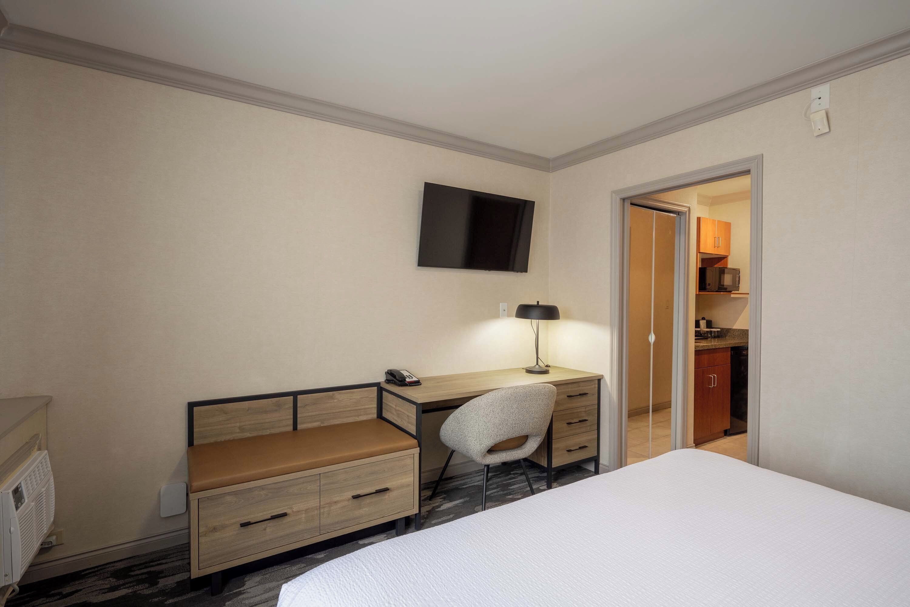 A hotel room with a bed, a desk with a chair, a flat-screen TV, and a kitchenette in the background.