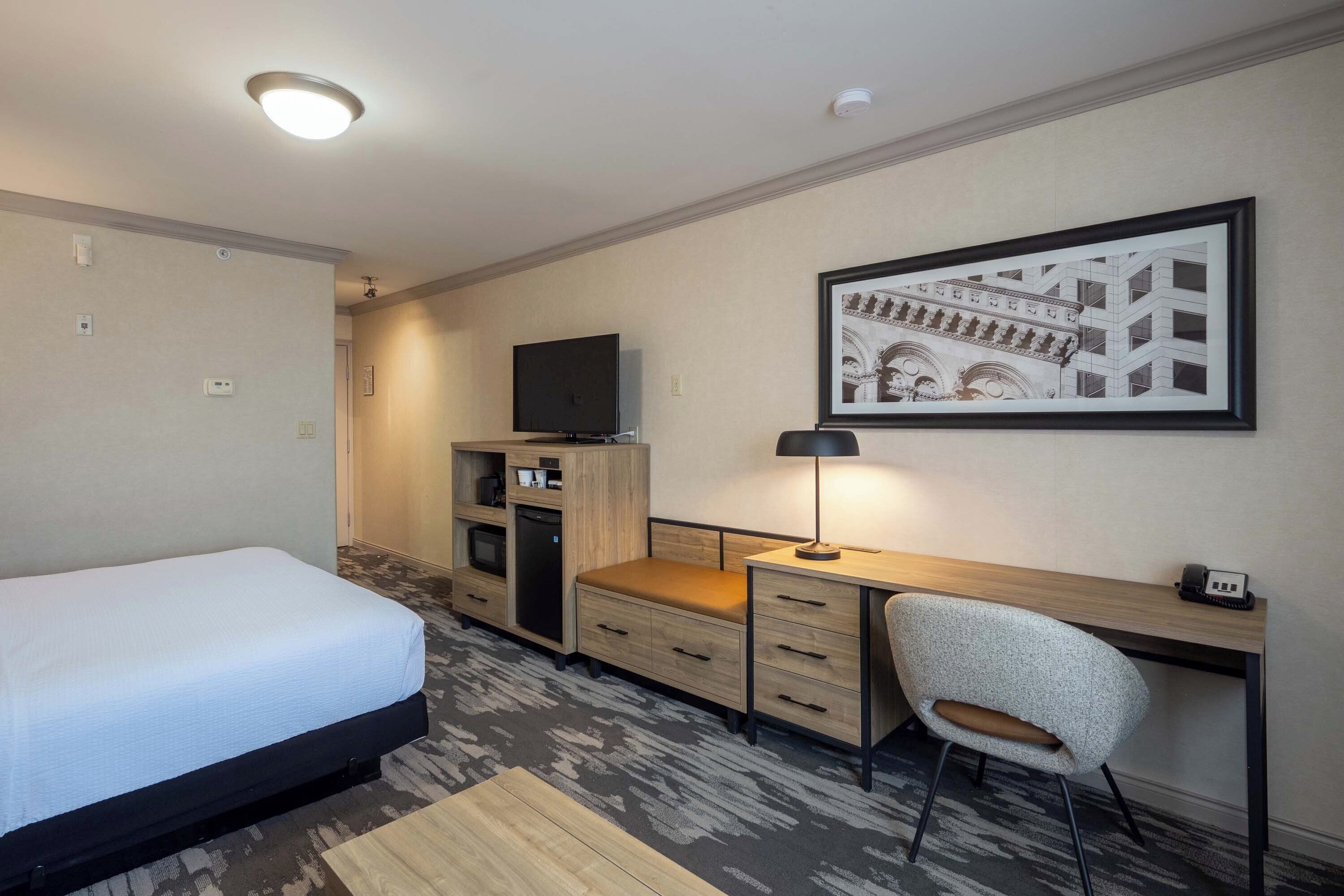A hotel room with a bed, desk, chair, TV, and a framed picture on the wall.