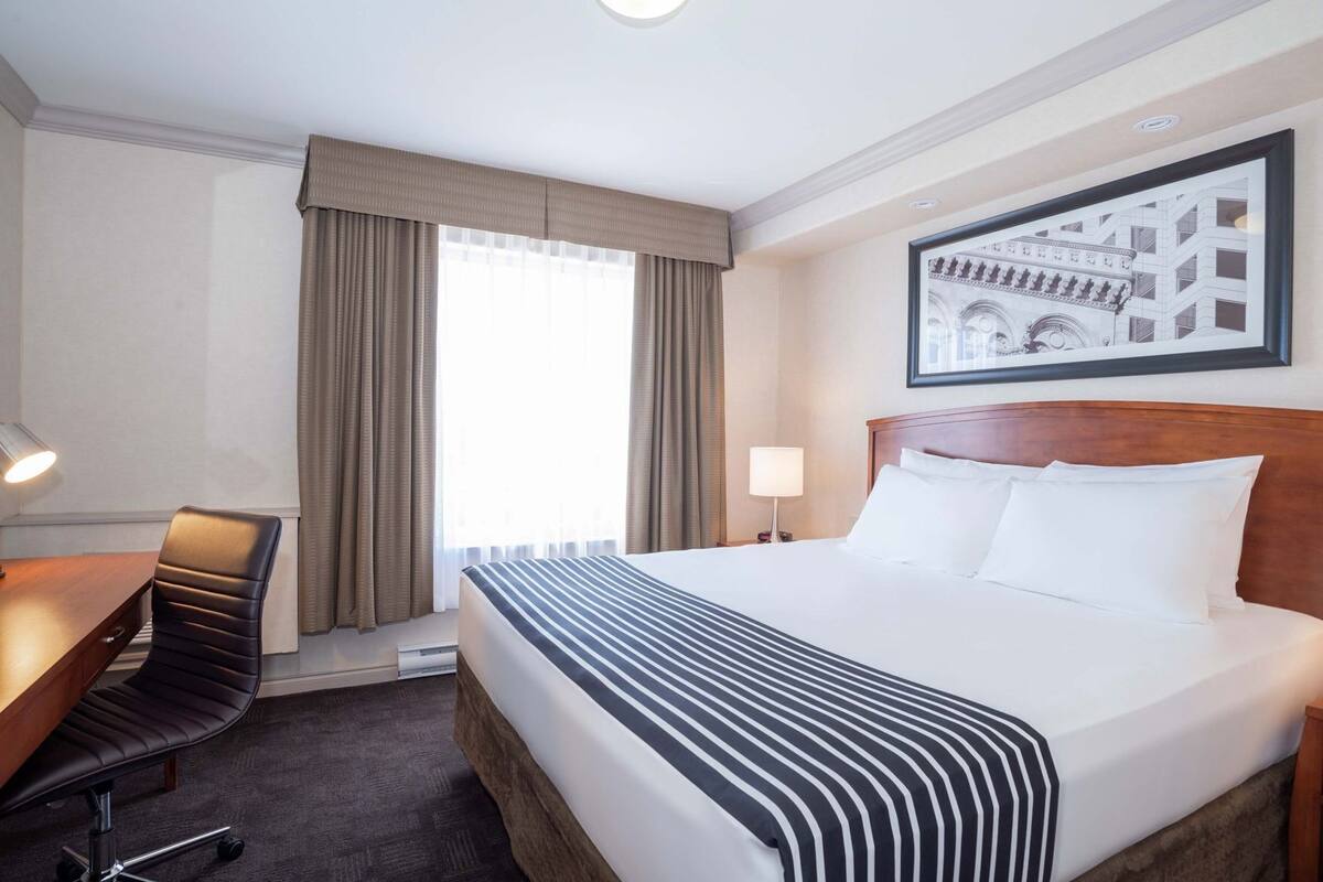 Une chambre d’hôtel avec un lit, un bureau, une chaise, une fenêtre avec des rideaux, une lampe et un tableau encadré au mur.