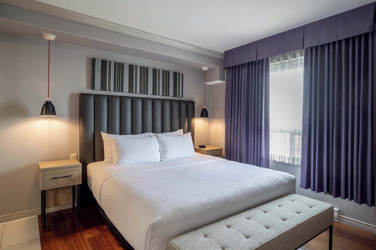 Une chambre d’hôtel moderne avec un grand lit, une tête de lit à rayures verticales, deux tables de chevet avec des lampes et une fenêtre avec des rideaux.