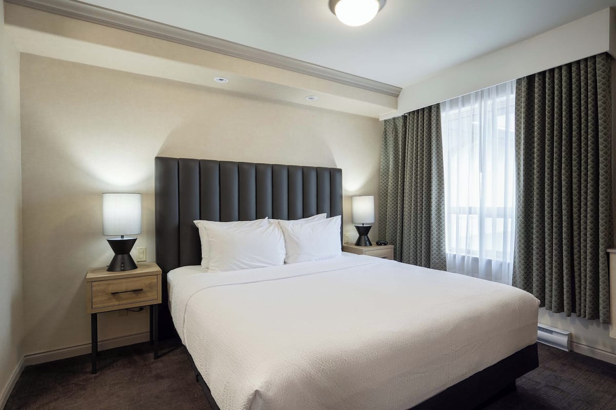 Une chambre d’hôtel avec un grand lit, deux tables de chevet avec des lampes, une tête de lit et une fenêtre avec des rideaux.