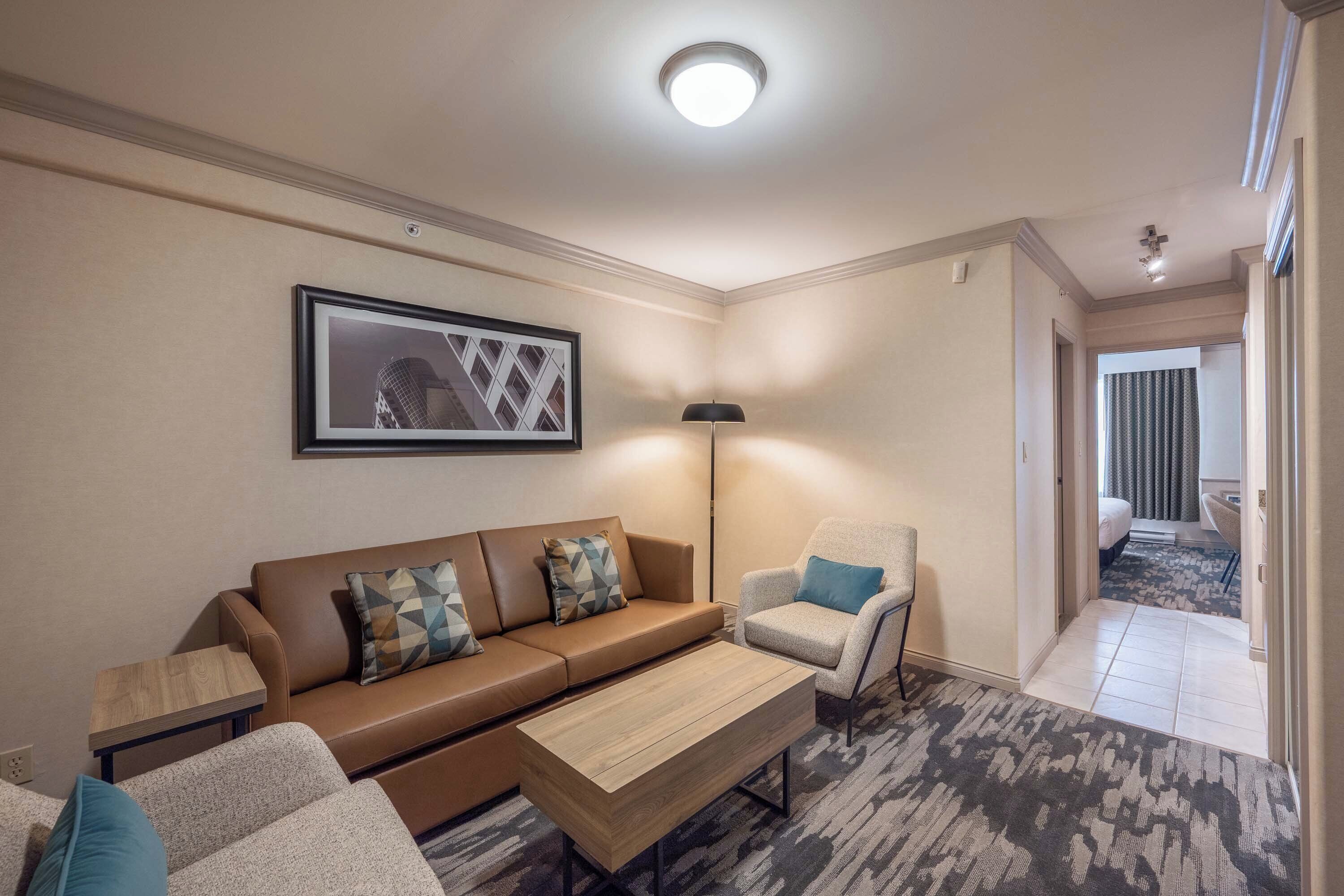 A modern living room with a brown sofa, a matching armchair, a coffee table, and a floor lamp. A framed picture hangs on the wall, and a carpeted floor covers the space.
