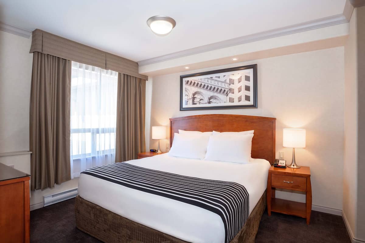 Une chambre d’hôtel avec un grand lit, deux tables de chevet avec des lampes, un tableau encadré au mur et une fenêtre avec des rideaux.