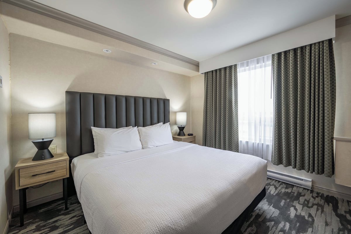 Une chambre d’hôtel avec un grand lit, deux tables de chevet avec des lampes, une tête de lit, une fenêtre avec des rideaux et un sol recouvert de moquette.