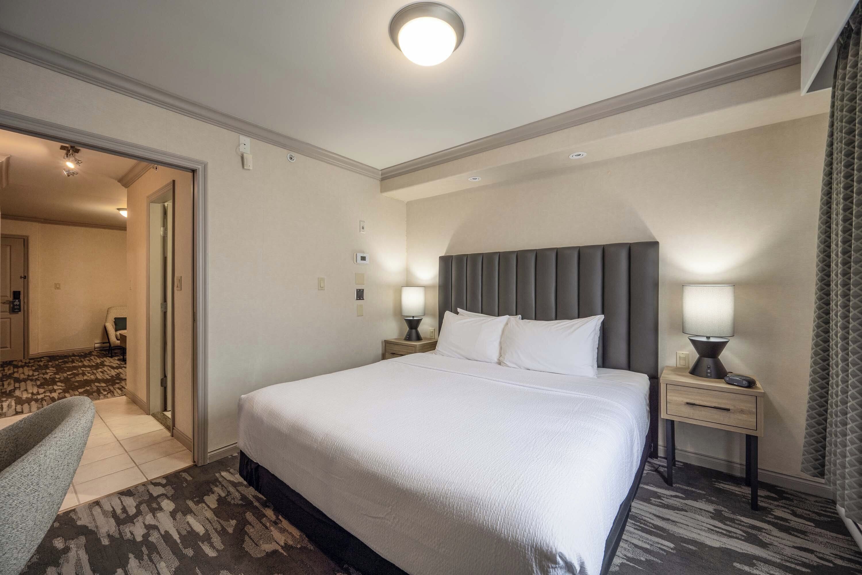 Una habitación de hotel con una cama grande, dos mesitas de noche con lámparas, una silla y suelo alfombrado.