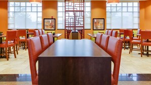 A spacious dining area with a long table and multiple chairs, large windows, and orange walls.