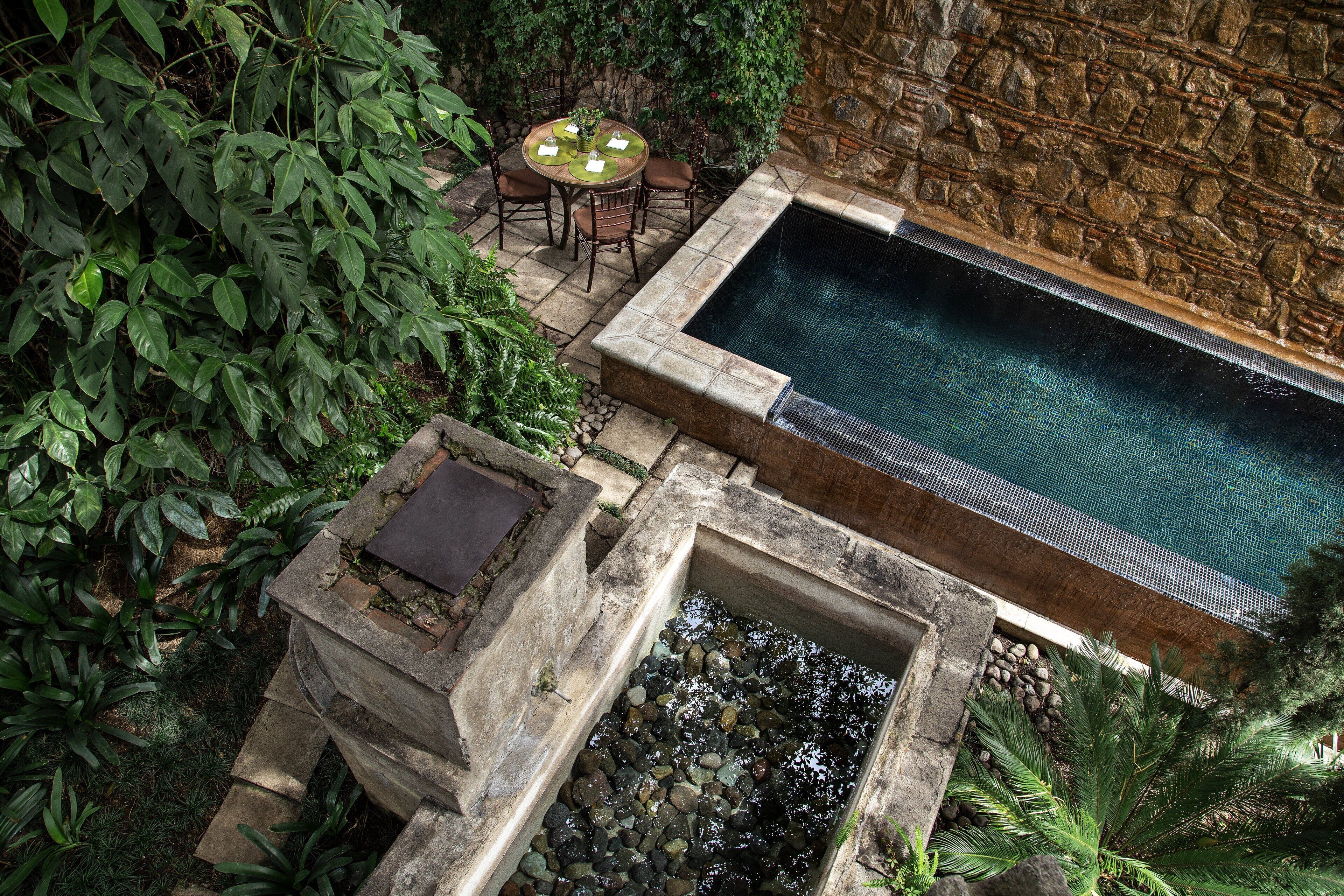 Una piscina de piedra rodeada de abundante vegetación y un muro de piedra.