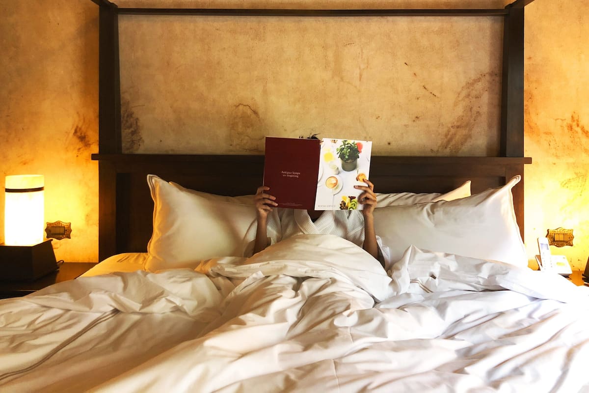 Una persona recostada en la cama leyendo un libro, en una habitación con cabecera de madera y lámparas de noche.