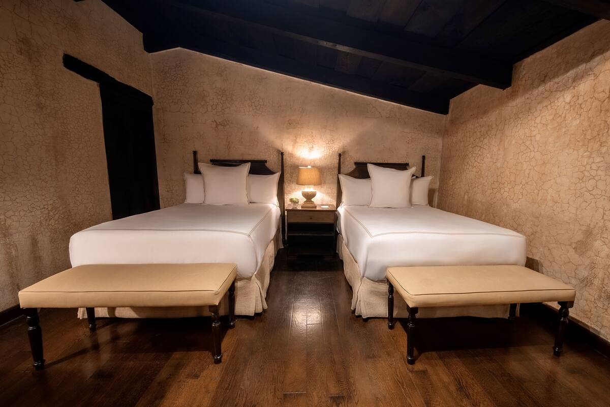 Una habitación con dos camas, un banco y una mesita de noche con lámpara.