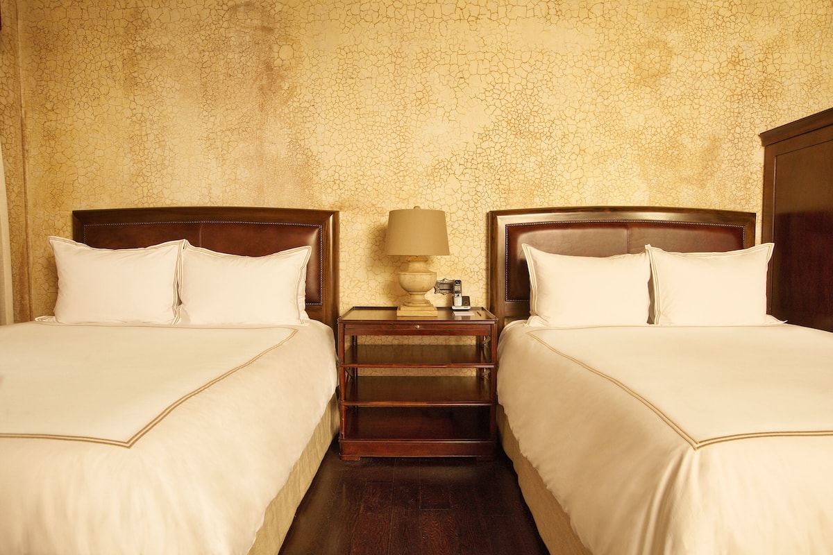 Habitación de hotel con dos camas, una mesita de noche con lámpara y pared con textura.