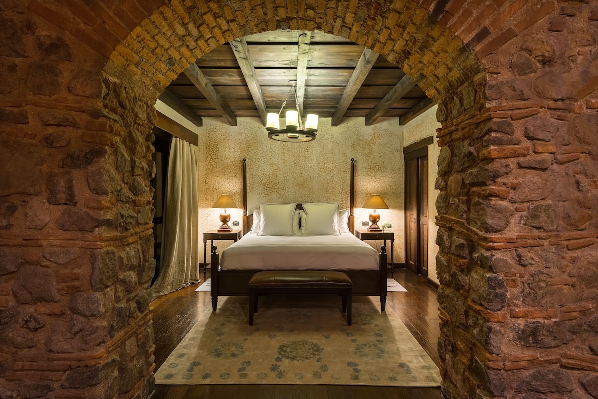 Un dormitorio con un arco de piedra, techo de madera, cama con cabecera, dos mesitas de noche con lámparas y un banco.