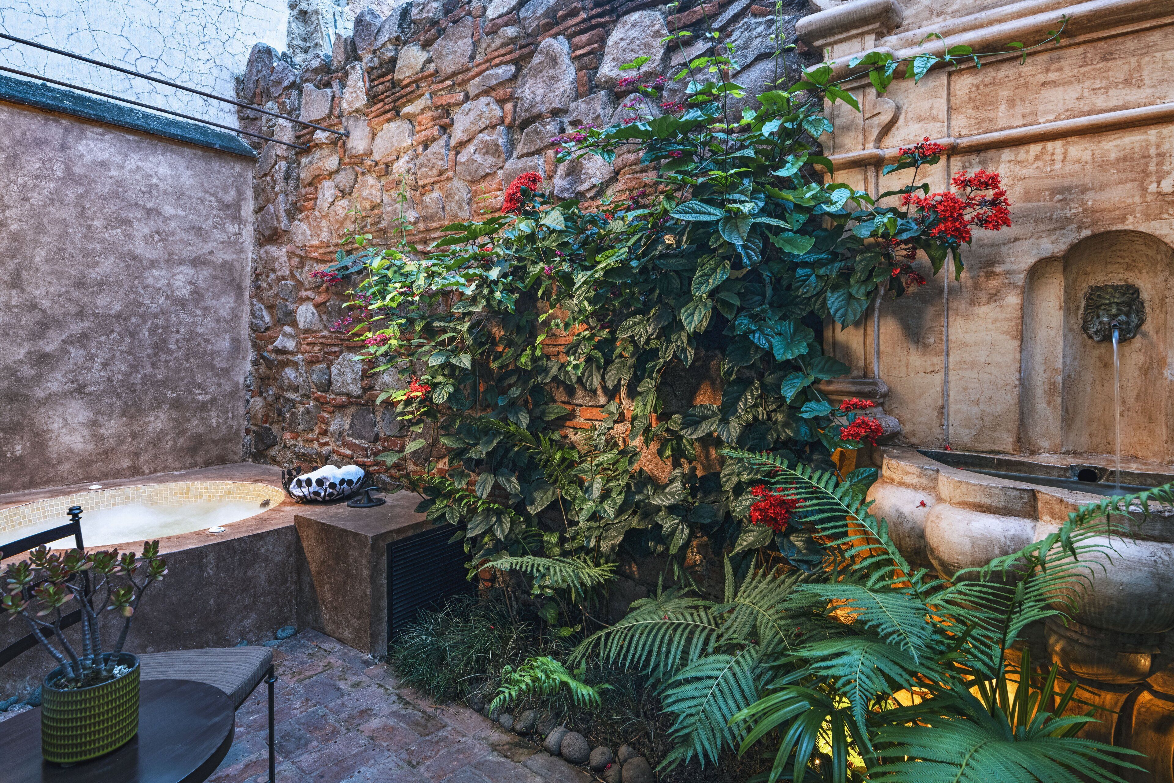 Zona de spa con paredes de piedra, bañera de hidromasaje, abundante vegetación y una fuente decorativa.