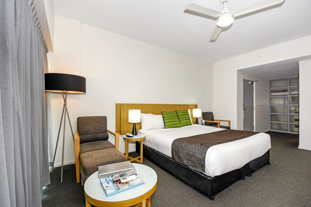 A hotel room with a bed, two bedside tables, a chair, a desk, a lamp, and a ceiling fan.