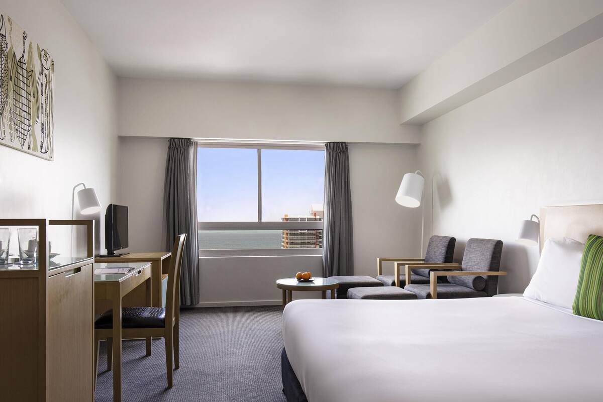 A hotel room with a bed, desk, chair, and a view of the sea.