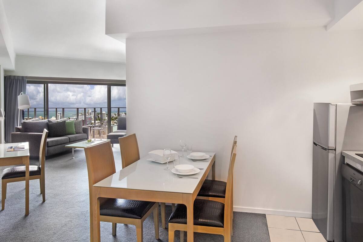 A modern hotel room with a dining area, a sofa, a balcony with a city view, and a kitchen area.