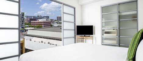 A hotel room with a bed, a TV on a stand, and large windows offering a city view.