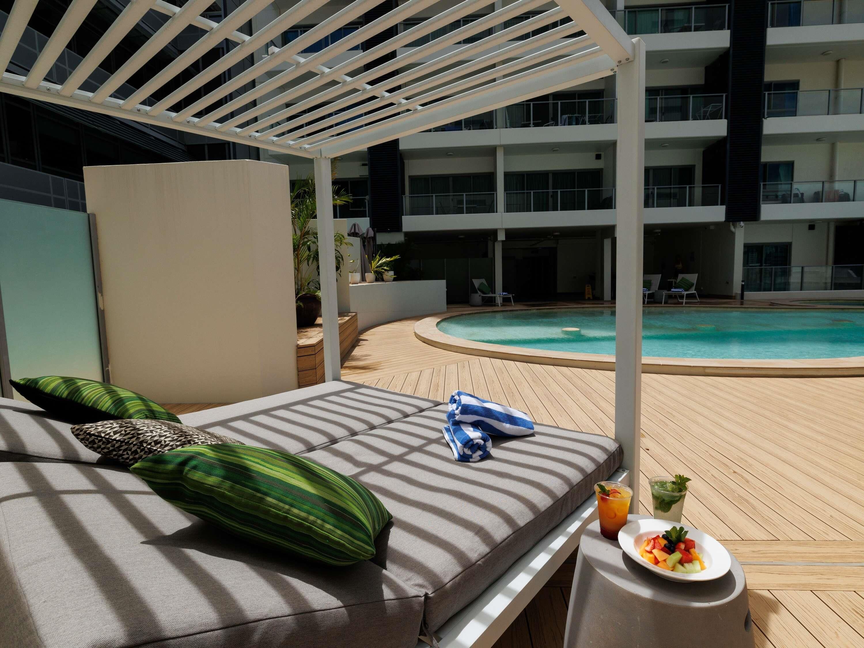 Ein Pool-Lounge-Bereich mit Liege, Tisch, Getränken und Früchten, mit Blick auf den Pool und das Gebäude im Hintergrund.