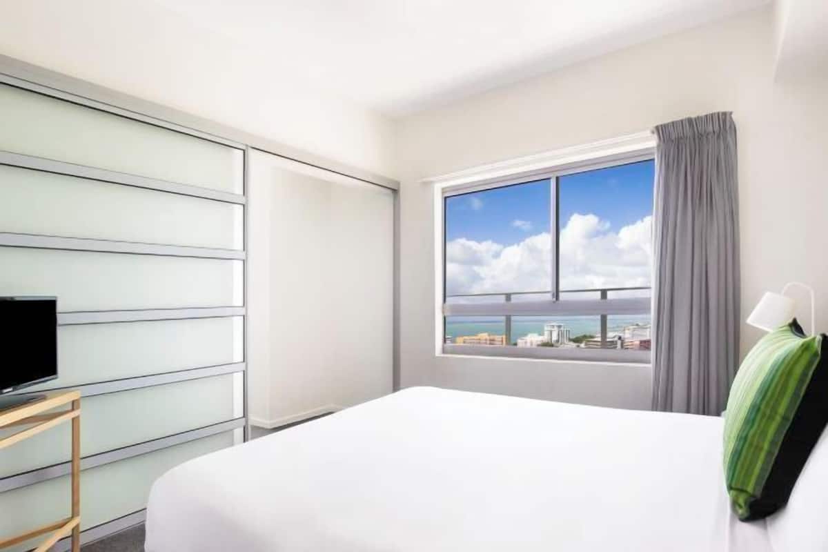 A hotel room with a bed, a TV on a stand, a window with a city view, and a curtain.