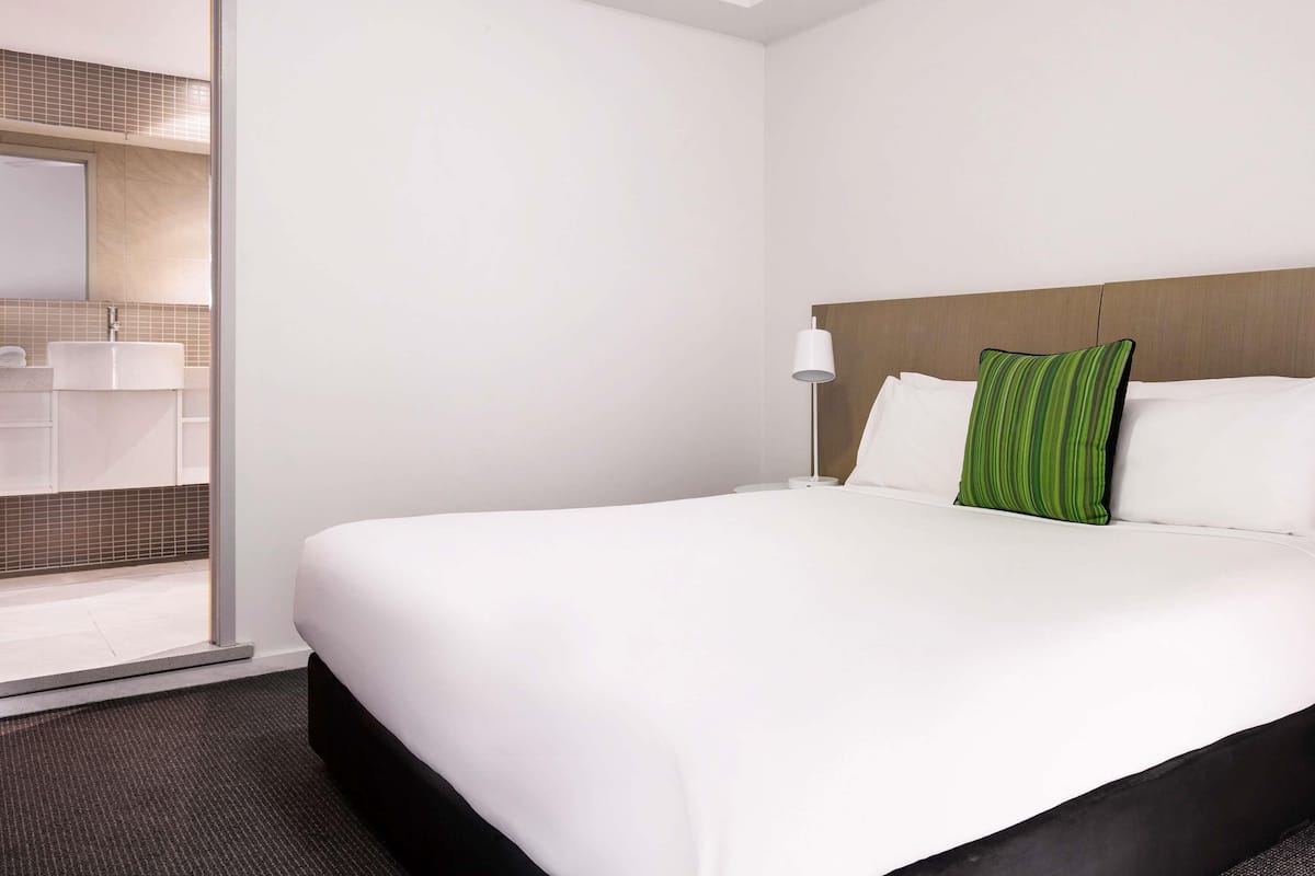A hotel room with a bed, a nightstand, a lamp, a green striped pillow, and a bathroom visible through an open door.