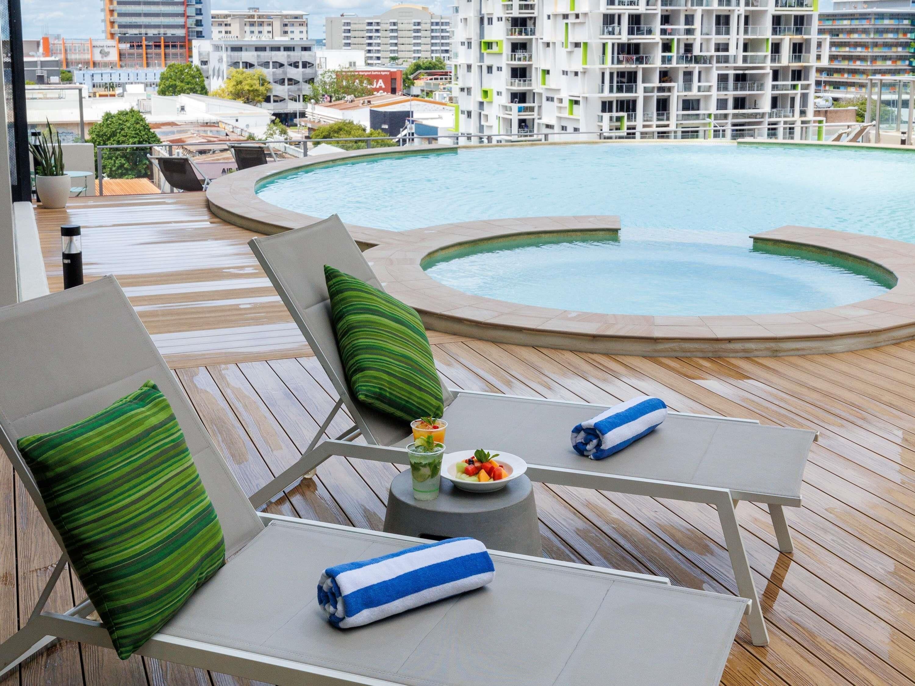A poolside area with lounge chairs, a small table with a drink and fruit, and a view of a cityscape with modern buildings.