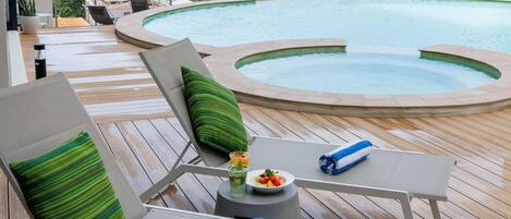 Zona de piscina con sillas de descanso, una pequeña mesa con una bebida y fruta, y vista a un paisaje urbano con edificios modernos.