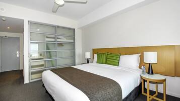 A hotel room with a large bed, a wooden headboard, two bedside tables with lamps, a ceiling fan, and a sliding glass door leading to a balcony.