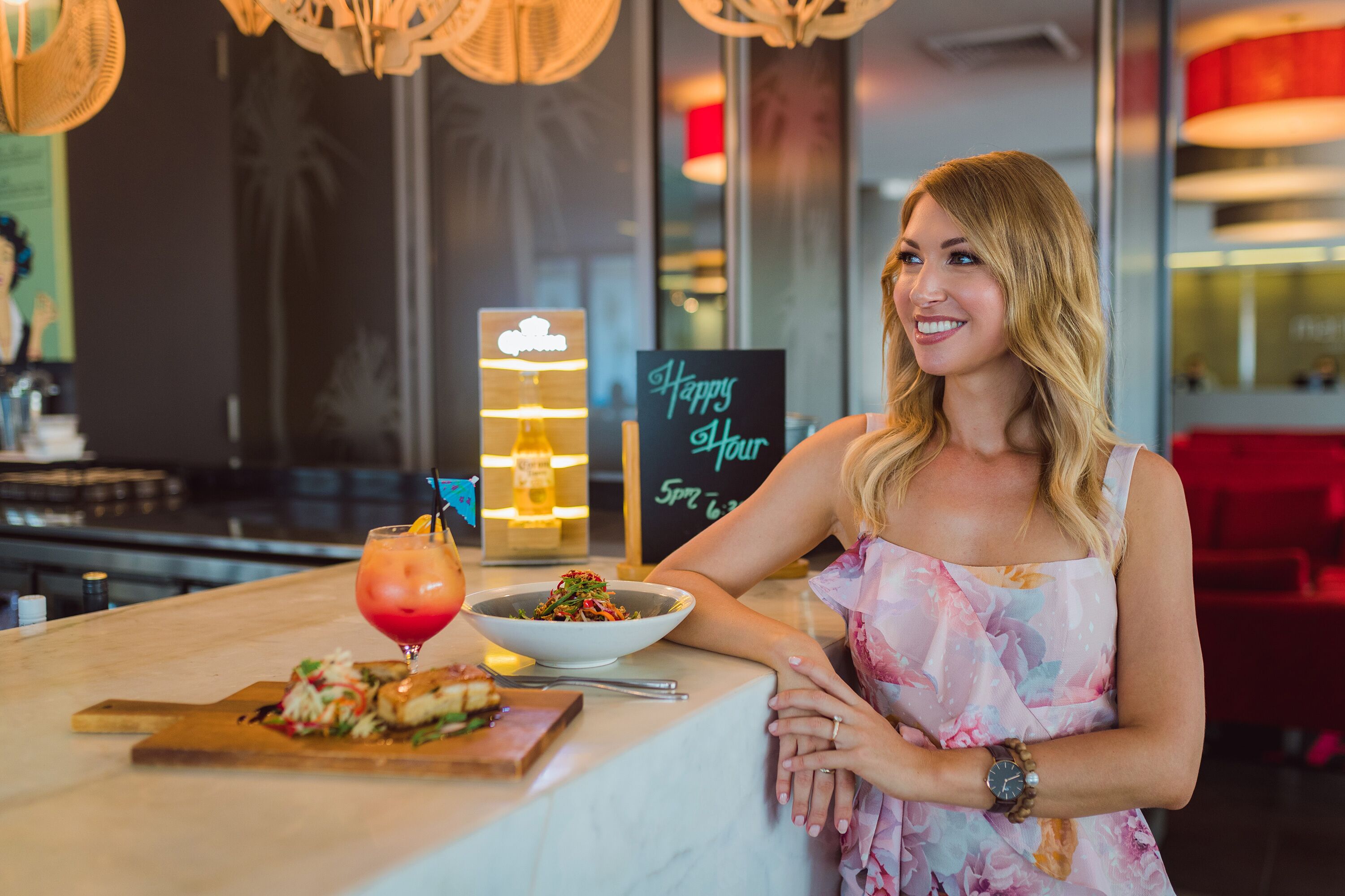 Une femme est assise au bar, devant un verre et un plat de nourriture. On peut voir une pancarte indiquant « Happy Hour » et une autre indiquant « de 17 h à 5 h du matin ».