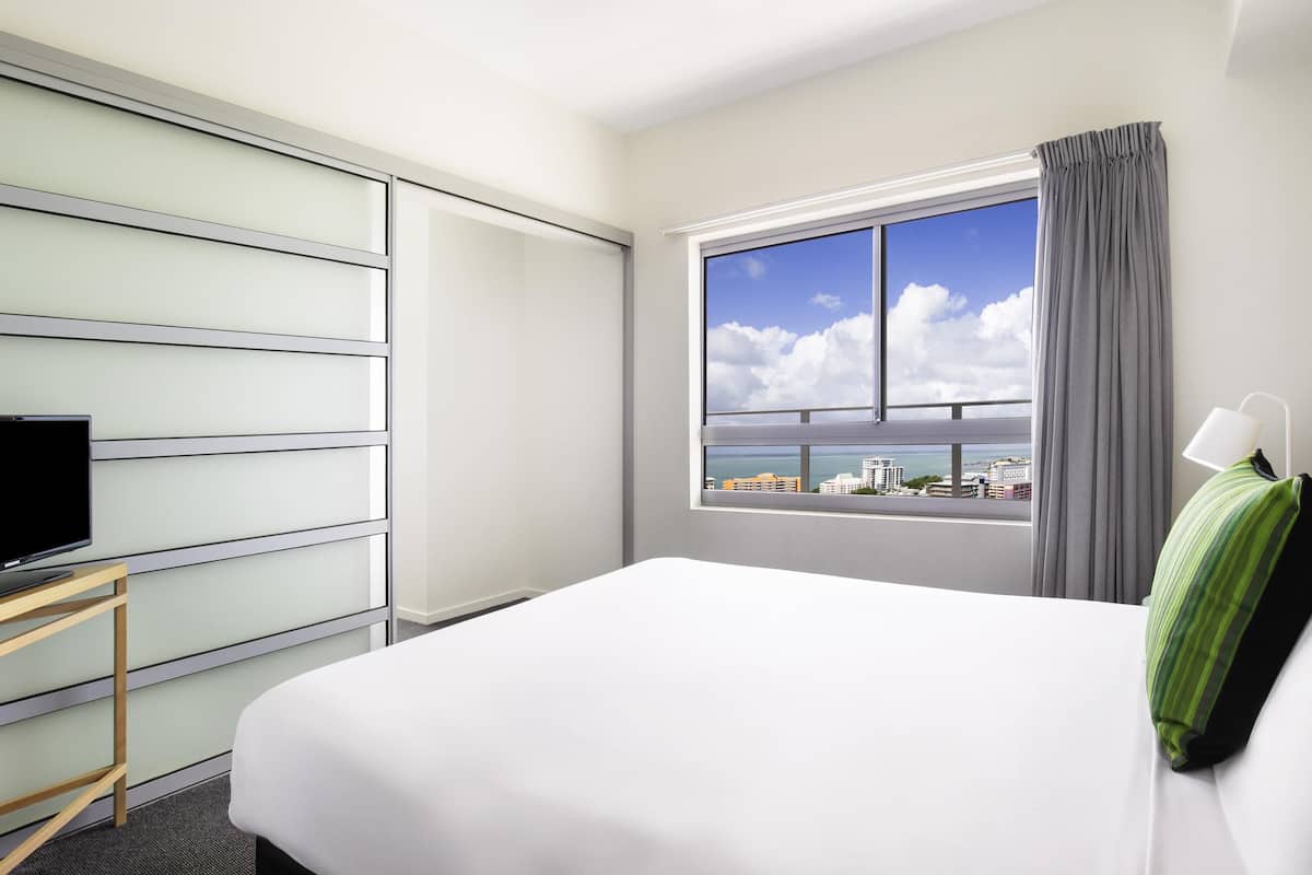 A hotel room with a bed, a TV on a stand, a window with a view of buildings and the sea, and a curtain.