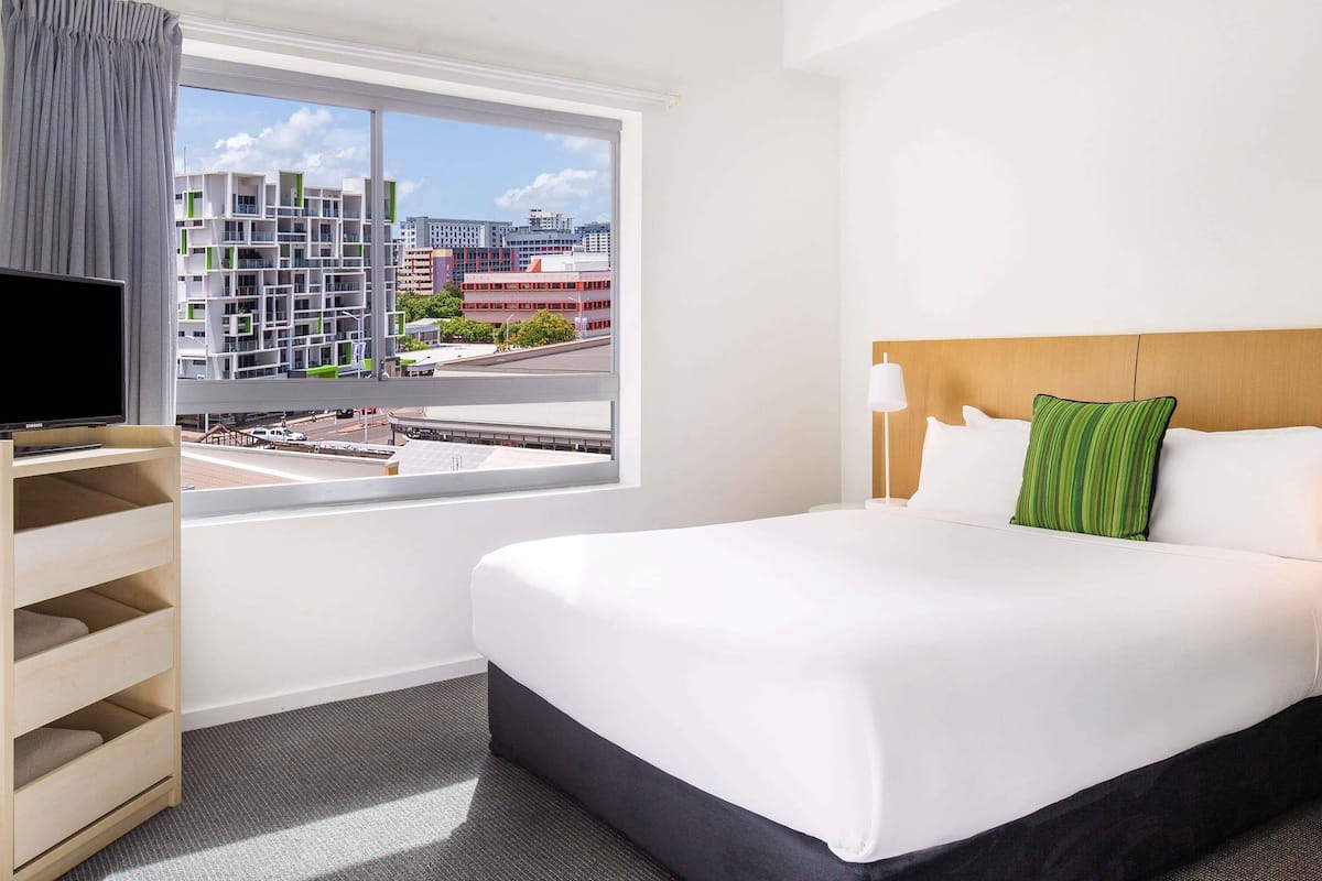 A hotel room with a bed, a TV on a stand, a window with a view of buildings, and a wooden shelf with drawers.