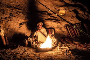 Une personne assise près d’un feu de camp dans un endroit qui ressemble à une grotte, avec un intérieur rustique.