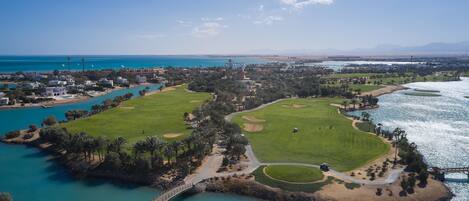 An aerial view of a coastal golf course with a bridge, water bodies, and surrounding greenery.