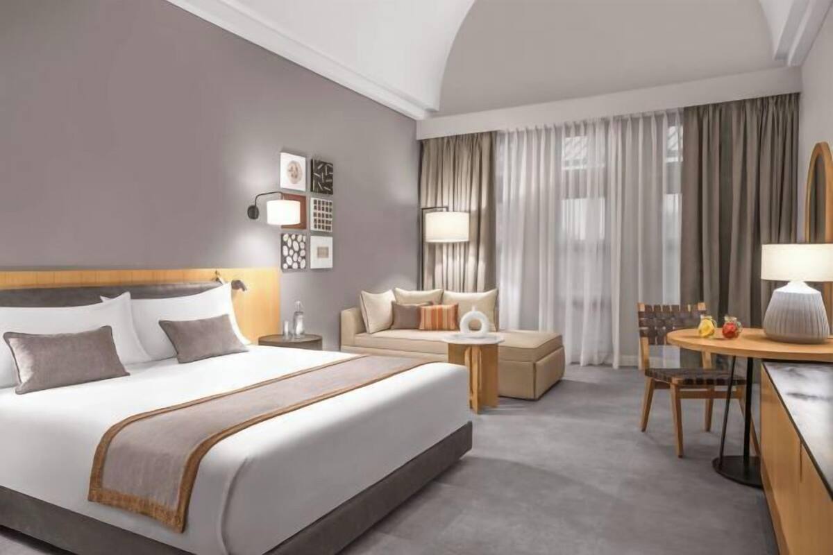 A hotel room with a large bed, a sofa, a small table, and a chair. There is a lamp on the table and a lamp on the side of the bed.