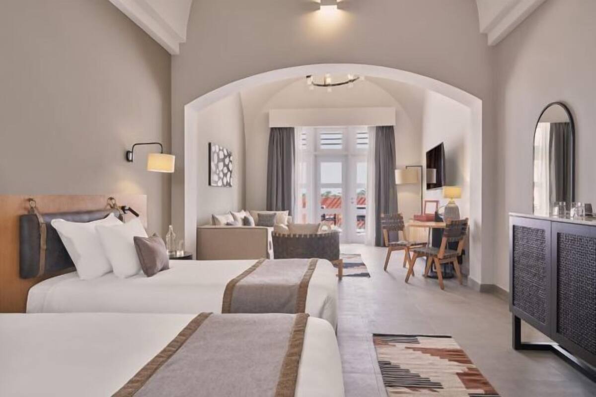 A hotel room with two beds, a desk, a chair, a television, and a view through an arched doorway.