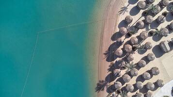 Vista aérea de una playa con aguas turquesas cristalinas, una orilla arenosa y hileras de sombrillas de paja.