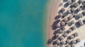 Vista aerea di una spiaggia con acque cristalline turchesi, una riva sabbiosa e file di ombrelloni di paglia.