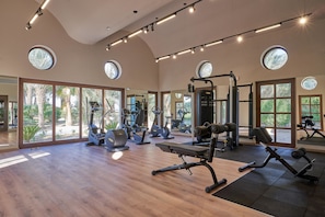 Een ruime fitnessruimte met houten vloeren, diverse fitnessapparaten en grote ramen die natuurlijk licht en uitzicht op groen bieden.