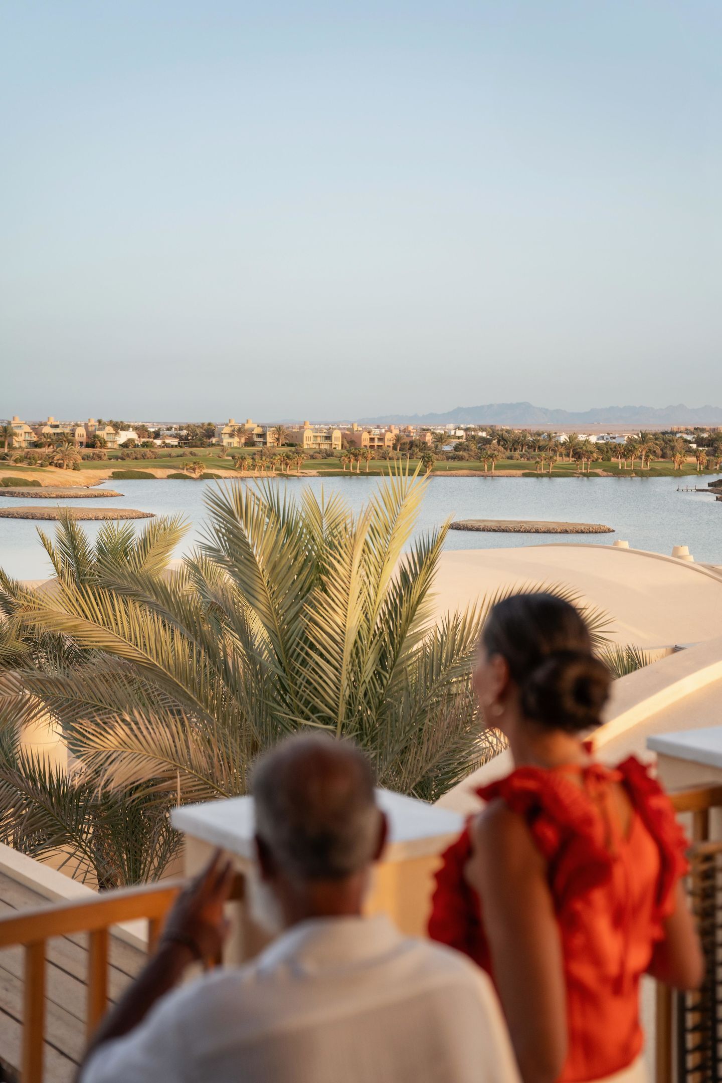 A balcony overlooking a body of water with palm trees, a sandy beach, and a distant cityscape.