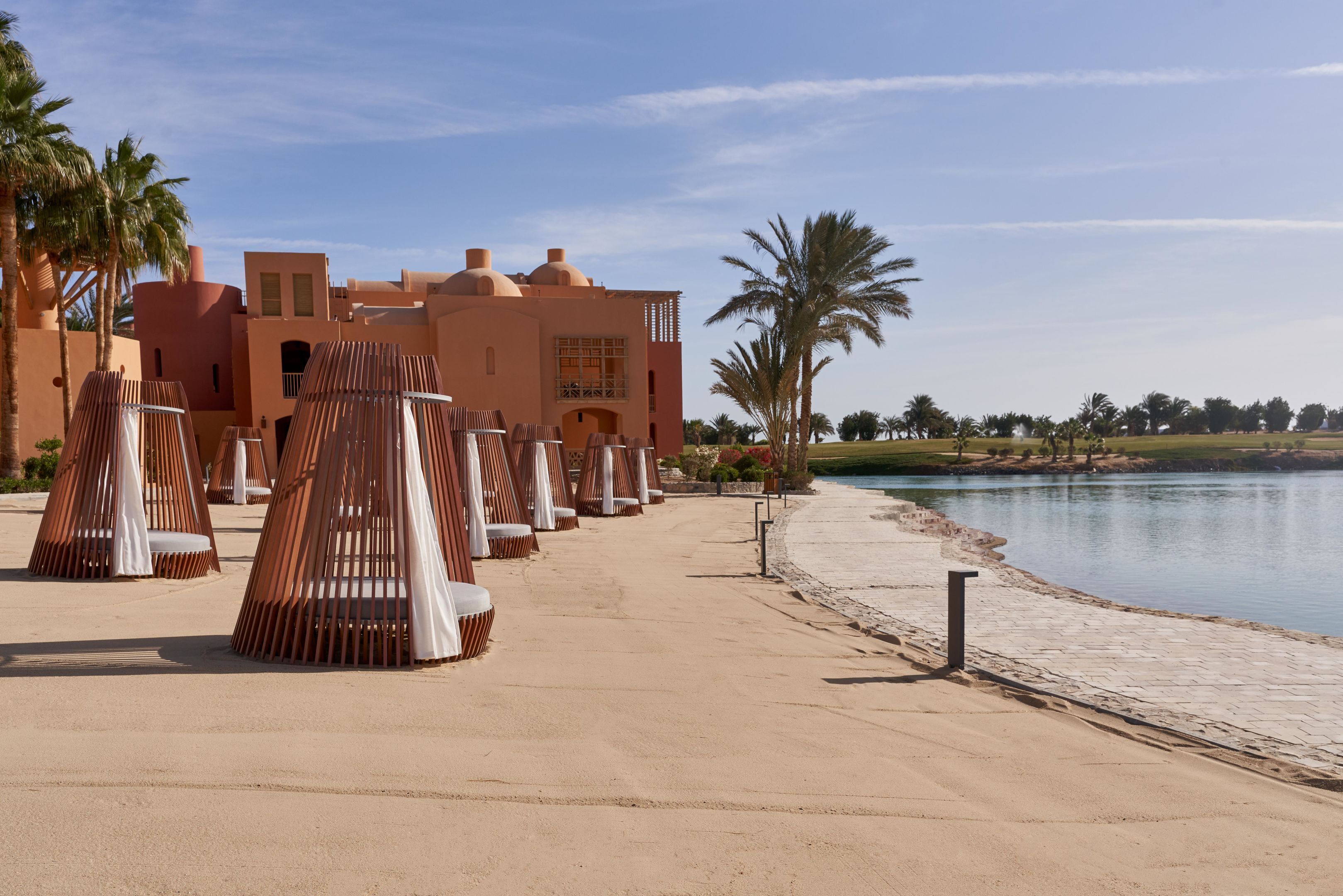 Un resort con mesas al aire libre cerca de un lago, palmeras y un edificio de estilo arquitectónico singular.