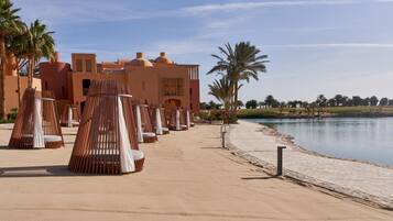 Un resort con mesas al aire libre cerca de un lago, palmeras y un edificio de estilo arquitectónico singular.