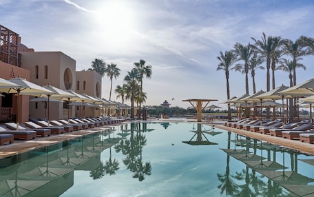 A poolside area with lounge chairs and umbrellas, surrounded by palm trees and a clear sky.