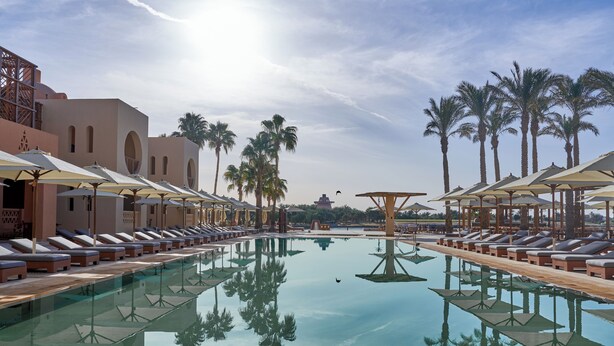 A poolside area with lounge chairs and umbrellas, surrounded by palm trees and a clear sky.